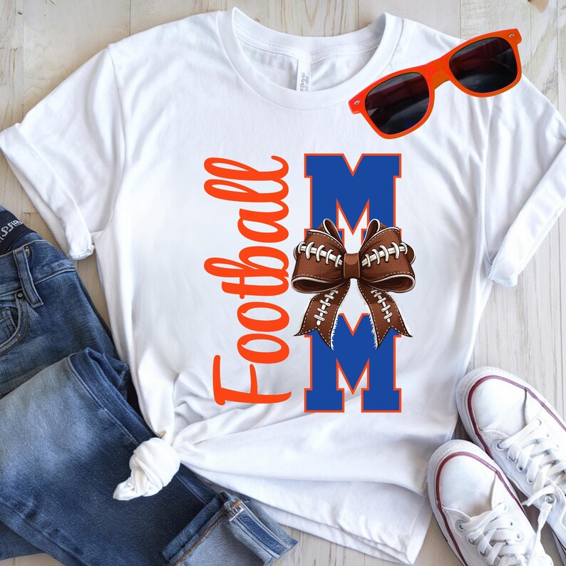 May include: White t-shirt with the text "Football Mom" in orange script. The word "MOM" is in blue block letters with a brown football-shaped bow in the center. Orange sunglasses rest on the shirt. Blue jeans and white sneakers are also visible.