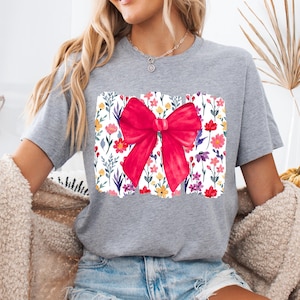 Peut inclure: Un t-shirt gris avec un motif de n&oelig;ud rouge vif. Le n&oelig;ud est centr&eacute; sur un motif floral rectangulaire avec des fleurs color&eacute;es. Le t-shirt est de style d&eacute;contract&eacute;, adapt&eacute; au quotidien.