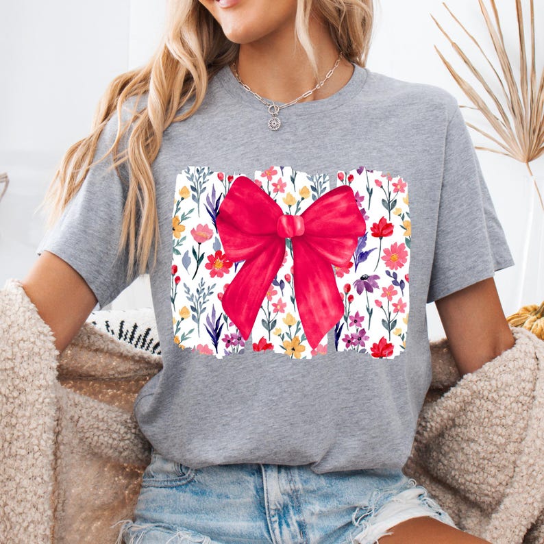Peut inclure: Un t-shirt gris avec un motif de n&oelig;ud rouge vif. Le n&oelig;ud est centr&eacute; sur un motif floral rectangulaire avec des fleurs color&eacute;es. Le t-shirt est de style d&eacute;contract&eacute;, adapt&eacute; au quotidien.