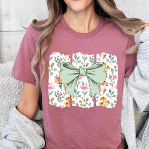 Op de afbeelding: Een mauvekleurig T-shirt met een groot, lichtgroen strikdesign. De strik is gecentreerd op een rechthoekig bloemenpatroon met roze, oranje en groene aquarelbloemen en -bladeren. Het T-shirt heeft korte mouwen.