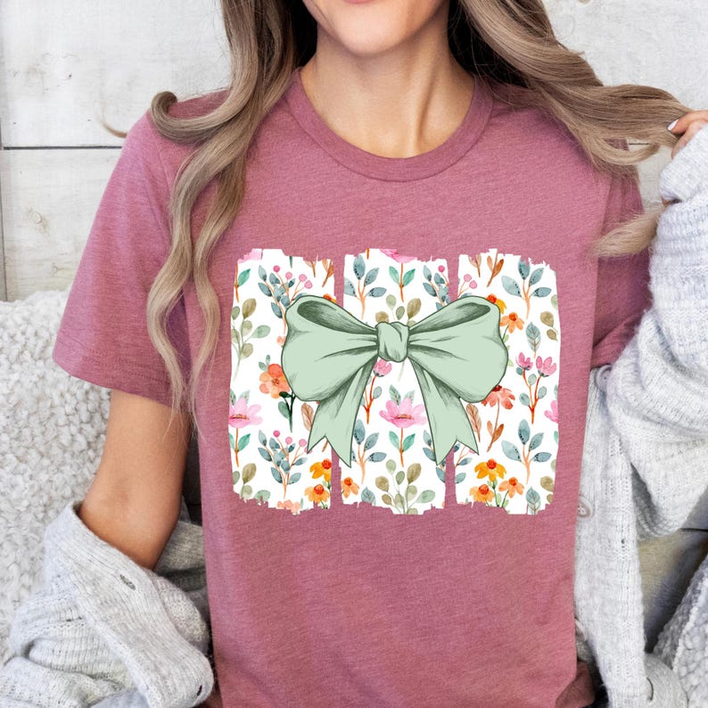 Op de afbeelding: Een mauvekleurig T-shirt met een groot, lichtgroen strikdesign. De strik is gecentreerd op een rechthoekig bloemenpatroon met roze, oranje en groene aquarelbloemen en -bladeren. Het T-shirt heeft korte mouwen.