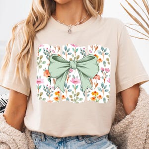 Op de afbeelding: Beige T-shirt met een aquarelontwerp van een grote, lichtgroene strik. De strik staat boven een rechthoekig bloemenpatroon met roze, oranje en blauwe bloemen en groene bladeren. Het shirt heeft korte mouwen.