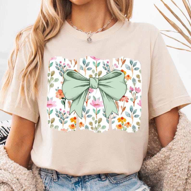 Op de afbeelding: Beige T-shirt met een aquarelontwerp van een grote, lichtgroene strik. De strik staat boven een rechthoekig bloemenpatroon met roze, oranje en blauwe bloemen en groene bladeren. Het shirt heeft korte mouwen.