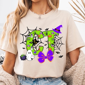 May include: A beige short-sleeve t-shirt featuring a graphic of the letters CT in bright green with a polka dot pattern. The design includes black spiderwebs, a purple witch hat, a white ghost, a purple cauldron, and a purple bow with orange trim.