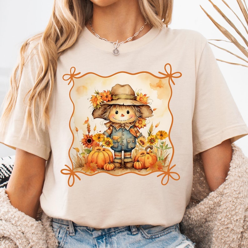 May include: Beige t-shirt featuring a watercolor illustration of a scarecrow wearing a hat adorned with sunflowers. The scarecrow is surrounded by pumpkins and fall foliage, set within an orange decorative frame. A perfect autumn-themed graphic tee.