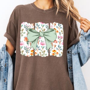 Op de afbeelding: Bruin t-shirt met een bloemenpatroon en een grote, lichtgroene strik. Het bloemenpatroon bevat roze, oranje en gele bloemen met groene bladeren op een witte achtergrond. Het shirt heeft een relaxte pasvorm.