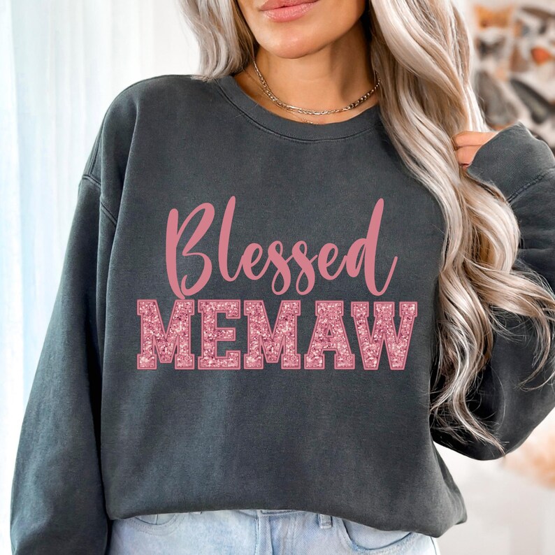 K&ouml;nnte beinhalten: Ein dunkelgrauer Pullover mit dem Schriftzug "Blessed Memaw" in rosafarbenen Glitzerbuchstaben.