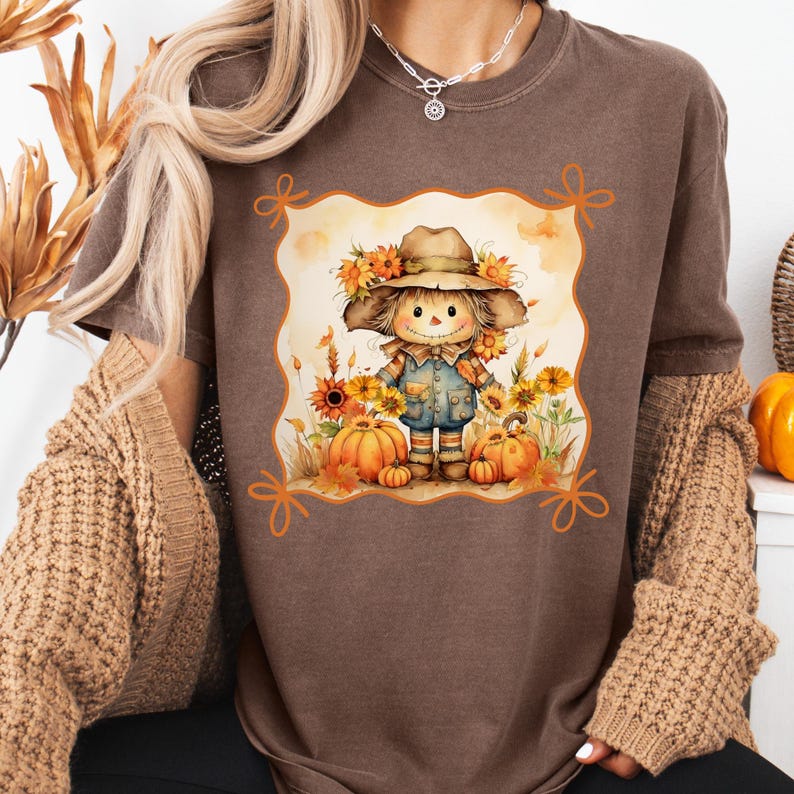 May include: Brown t-shirt featuring a framed illustration of a scarecrow with a hat, surrounded by sunflowers and pumpkins. The scarecrow wears overalls and stands in a field. The shirt is a solid brown color.