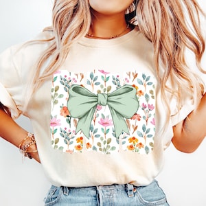 Op de afbeelding: Cr&egrave;mekleurig T-shirt met een groot, lichtgroen strikdesign. De strik is gecentreerd op een vierkant paneel met een bloemenpatroon van roze, oranje en blauwe bloemen en groene bladeren op een witte achtergrond.