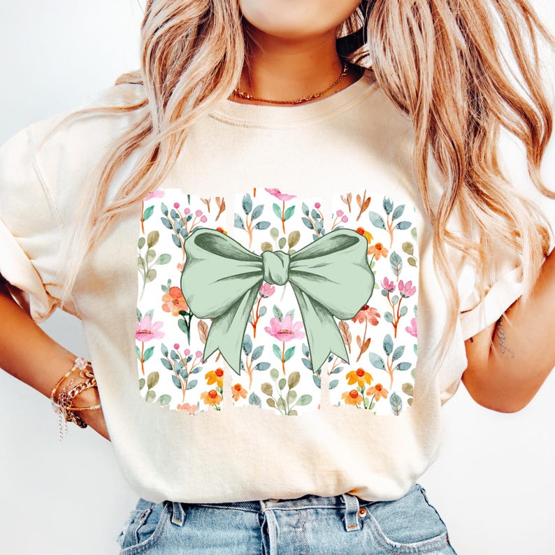 Op de afbeelding: Cr&egrave;mekleurig T-shirt met een groot, lichtgroen strikdesign. De strik is gecentreerd op een vierkant paneel met een bloemenpatroon van roze, oranje en blauwe bloemen en groene bladeren op een witte achtergrond.