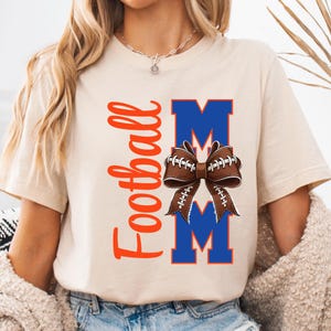 May include: A beige t-shirt with the words "Football Mom" in orange script. The design includes a brown football-shaped bow over two blue "M" letters. The shirt is a casual style.