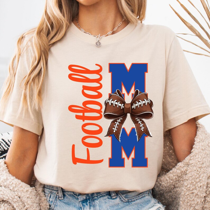May include: A beige t-shirt with the words "Football Mom" in orange script. The design includes a brown football-shaped bow over two blue "M" letters. The shirt is a casual style.
