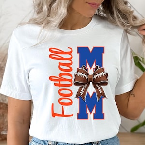 May include: White t-shirt with the words "Football Mom" in orange script and blue block letters. A brown football-shaped bow is centered between the letters "M" in the word "Mom".