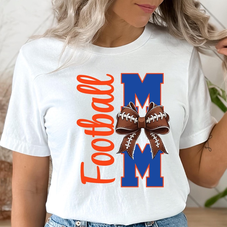 May include: White t-shirt with the words "Football Mom" in orange script and blue block letters. A brown football-shaped bow is centered between the letters "M" in the word "Mom".
