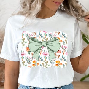 Op de afbeelding: Wit T-shirt met een grote, lichtgroene strik. De strik staat op een rechthoekig bloemenpatroon met roze, oranje en gele bloemen en groene bladeren. Een casual kledingstuk voor dagelijks gebruik.