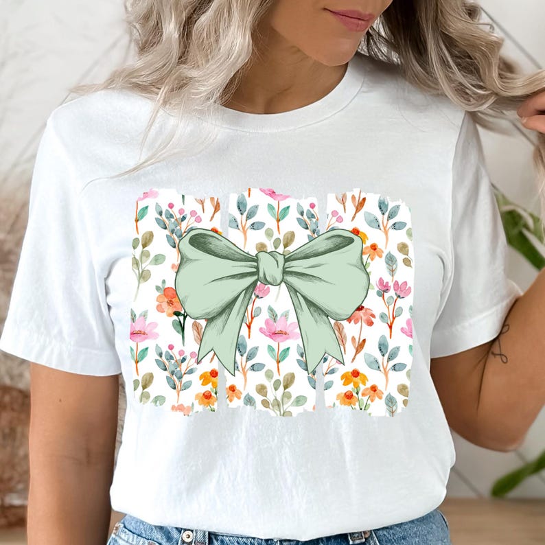 Op de afbeelding: Wit T-shirt met een grote, lichtgroene strik. De strik staat op een rechthoekig bloemenpatroon met roze, oranje en gele bloemen en groene bladeren. Een casual kledingstuk voor dagelijks gebruik.