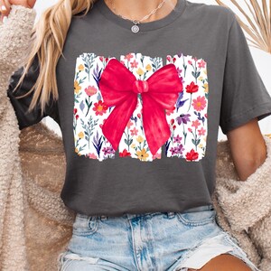 Peut inclure: T-shirt gris fonc&eacute; avec un grand n&oelig;ud rouge sur un motif floral. Le motif floral comprend des fleurs color&eacute;es et des feuilles vertes sur fond blanc. Le t-shirt est de style d&eacute;contract&eacute;.
