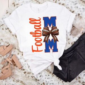 May include: White t-shirt with the words "Football Mom" in orange script and blue block letters. A brown football-shaped bow is centered between the letters "M". The shirt is folded and tied at the bottom.