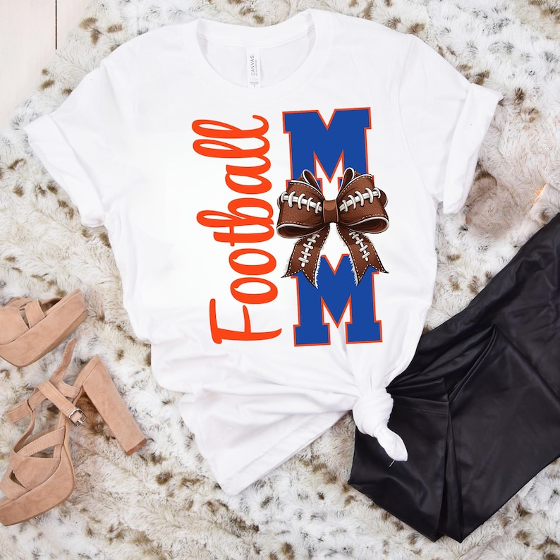 May include: White t-shirt with the words "Football Mom" in orange script and blue block letters. A brown football-shaped bow is centered between the letters "M". The shirt is folded and tied at the bottom.