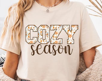 Puede incluir: Camiseta beige con las palabras "COZY season" en fuente marrón. Las letras de "COZY" están rellenas con un estampado floral de flores naranjas y blancas. La camiseta es de color neutro y tiene mangas cortas.