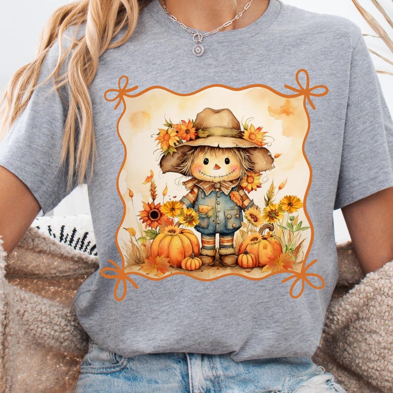 May include: A gray t-shirt featuring a watercolor illustration of a scarecrow with a hat, surrounded by sunflowers and pumpkins. The scarecrow wears overalls and a straw hat, with a decorative border.