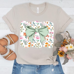 Op de afbeelding: Beige T-shirt met een groot, lichtgroen strikdesign over een bloemenpatroon. Het bloemenpatroon bevat roze, oranje en blauwe bloemen en bladeren. Het shirt wordt gedragen met een spijkerbroek en sandalen.