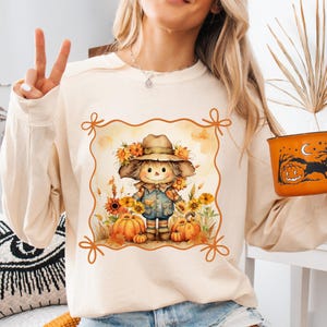May include: Cream-colored sweatshirt featuring a fall-themed graphic. The graphic depicts a scarecrow with sunflowers, pumpkins, and autumn leaves. The scarecrow wears a hat and overalls. An orange Halloween bucket is held in the hand.