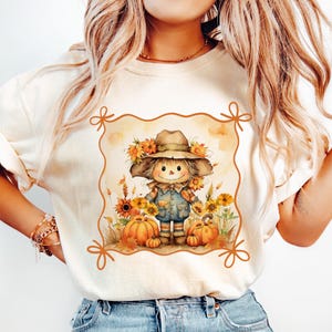May include: Cream-colored sweatshirt featuring a watercolor illustration of a scarecrow with a hat, surrounded by sunflowers and pumpkins. The design is framed with a decorative border, evoking a fall harvest theme.