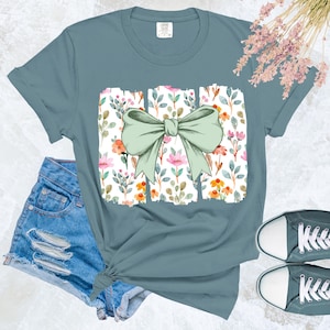 Op de afbeelding: Een lichtblauw T-shirt met een strikdesign en een bloemenpatroon. De strik heeft een zachte groene kleur en het bloemenpatroon bevat roze, oranje en witte bloemen. Het shirt wordt gecombineerd met een spijkerbroekshort en grijze canvas sneakers.