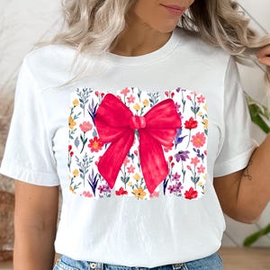 Peut inclure: T-shirt blanc avec un grand n&oelig;ud rouge vif. Le n&oelig;ud est plac&eacute; sur un motif floral rectangulaire avec des fleurs color&eacute;es et des feuilles vertes. Le t-shirt est en mati&egrave;re douce et confortable.
