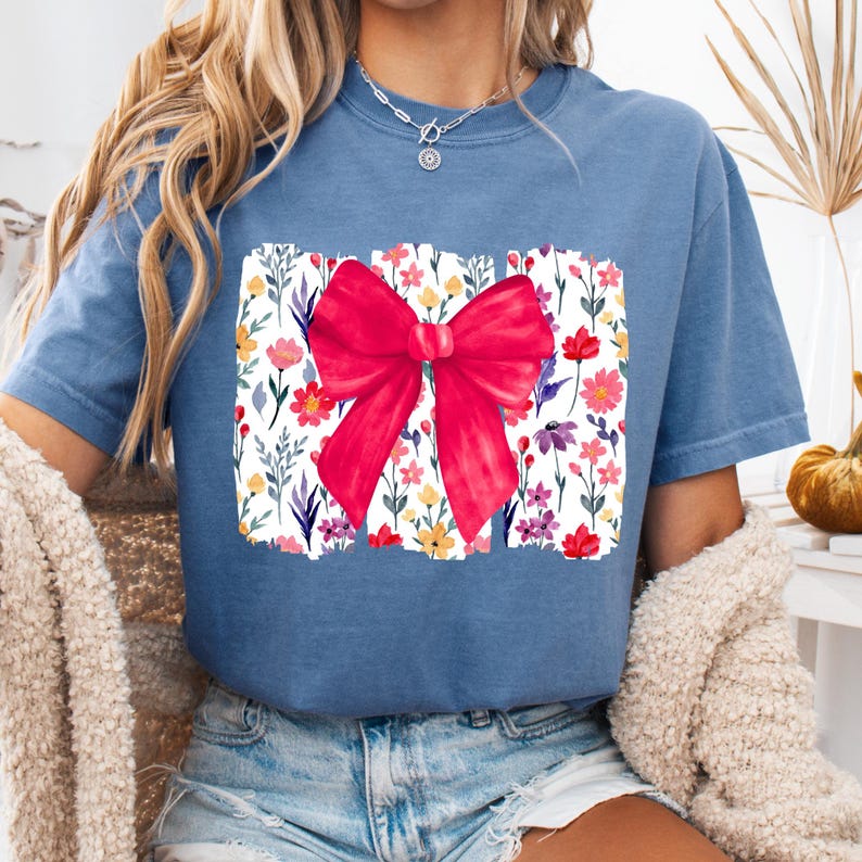Peut inclure: T-shirt bleu &agrave; manches courtes avec un grand n&oelig;ud rouge. Le n&oelig;ud est plac&eacute; sur un motif rectangulaire rempli de fleurs et de feuillages color&eacute;s. Le t-shirt est de couleur bleu uni.