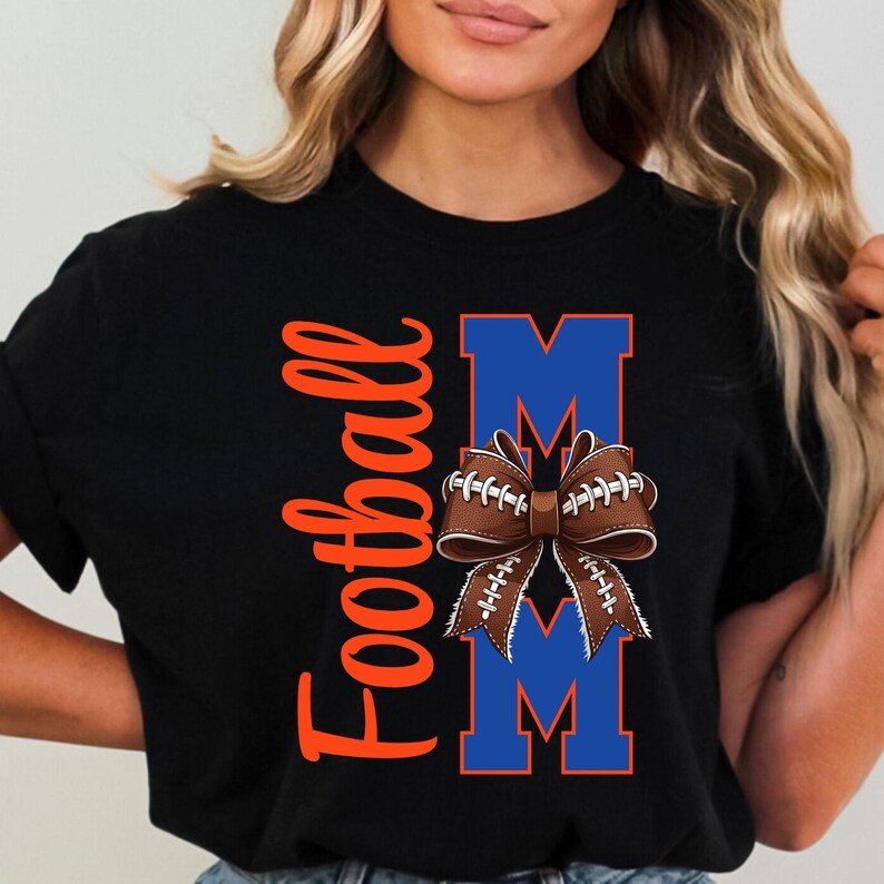 May include: Black t-shirt with the word "Football" in orange script and the word "MOM" in blue block letters. A brown football-shaped bow with white laces and blue accents is centered between the letters.