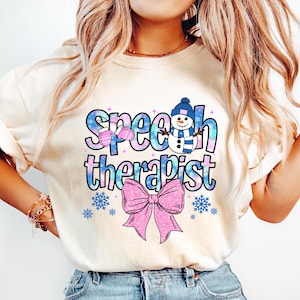 May include: Cream-colored sweatshirt with the words "Speech Therapist" in a blue and pink design. The design includes a snowman wearing a blue hat and scarf, pink mittens, snowflakes, and a large pink bow.