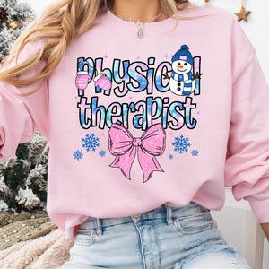 May include: Pink sweatshirt with the words "Physical Therapist" in a blue and white design, along with a snowman, mittens, snowflakes, and a pink bow. The sweatshirt is a crew neck style.