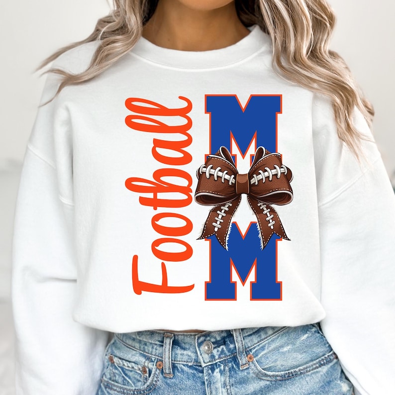 May include: White sweatshirt with the word "Football" in orange script on the left side. The word "MOM" is in blue block letters, with a football-shaped bow in the center. The bow is brown with white stitching.
