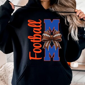 May include: Black hooded sweatshirt with the word "Football" in orange script. The word "MOM" is in blue block letters with a football-shaped bow in the center. The bow is brown with white laces.