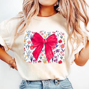 Peut inclure: Sweat-shirt cr&egrave;me avec un motif de n&oelig;ud rouge vif. Le n&oelig;ud est centr&eacute; sur un motif floral rectangulaire avec des fleurs color&eacute;es et des feuilles vertes. L'ensemble d&eacute;gage une esth&eacute;tique joyeuse et f&eacute;minine.