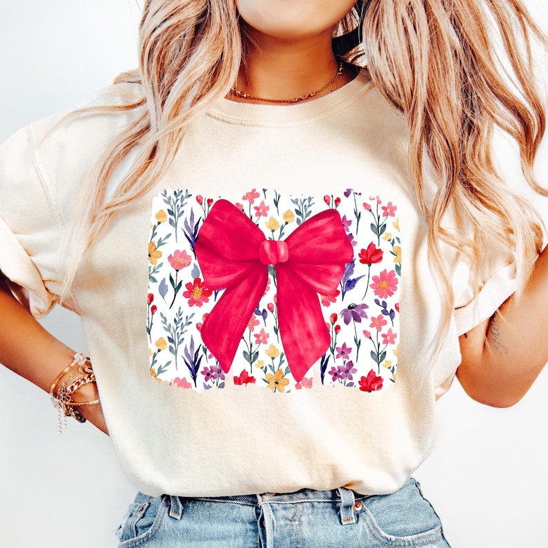 Peut inclure: Sweat-shirt cr&egrave;me avec un motif de n&oelig;ud rouge vif. Le n&oelig;ud est centr&eacute; sur un motif floral rectangulaire avec des fleurs color&eacute;es et des feuilles vertes. L'ensemble d&eacute;gage une esth&eacute;tique joyeuse et f&eacute;minine.