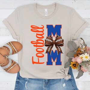 May include: A beige t-shirt with the words "Football Mom" in orange script and blue block letters. A football-shaped bow is centered between the letters "M". The shirt is paired with denim shorts and sandals.