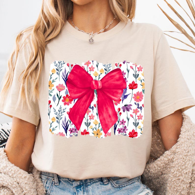 Peut inclure: T-shirt beige avec un grand n&oelig;ud rouge vif. Le n&oelig;ud est plac&eacute; sur un motif floral rectangulaire avec des fleurs aquarelles color&eacute;es. Le t-shirt a des manches courtes et un col rond.
