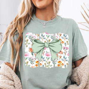 May include: A light green t-shirt featuring a large, light green bow over a floral pattern. The floral design includes pink, orange, and yellow flowers with green leaves on a white background. The shirt has short sleeves.