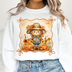 May include: White sweatshirt featuring a watercolor illustration of a scarecrow wearing a hat adorned with sunflowers. The scarecrow is surrounded by pumpkins and more sunflowers, with an orange border.