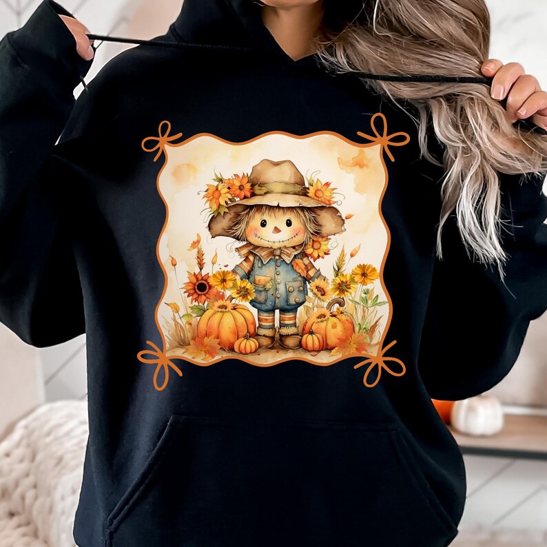 May include: Black hooded sweatshirt featuring a fall-themed graphic. The graphic depicts a smiling scarecrow wearing a hat and surrounded by sunflowers and pumpkins. The design is framed by decorative orange bows.