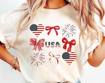 May include: Cream-colored sweatshirt featuring patriotic designs. The design includes hearts with American flags, red bows, fireworks, and the word "USA" in red and blue. A festive and casual top.
