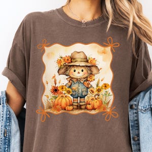 May include: Brown t-shirt featuring a fall-themed graphic. The graphic depicts a smiling scarecrow wearing a hat and overalls, surrounded by pumpkins and sunflowers. The design is framed with orange decorative elements.