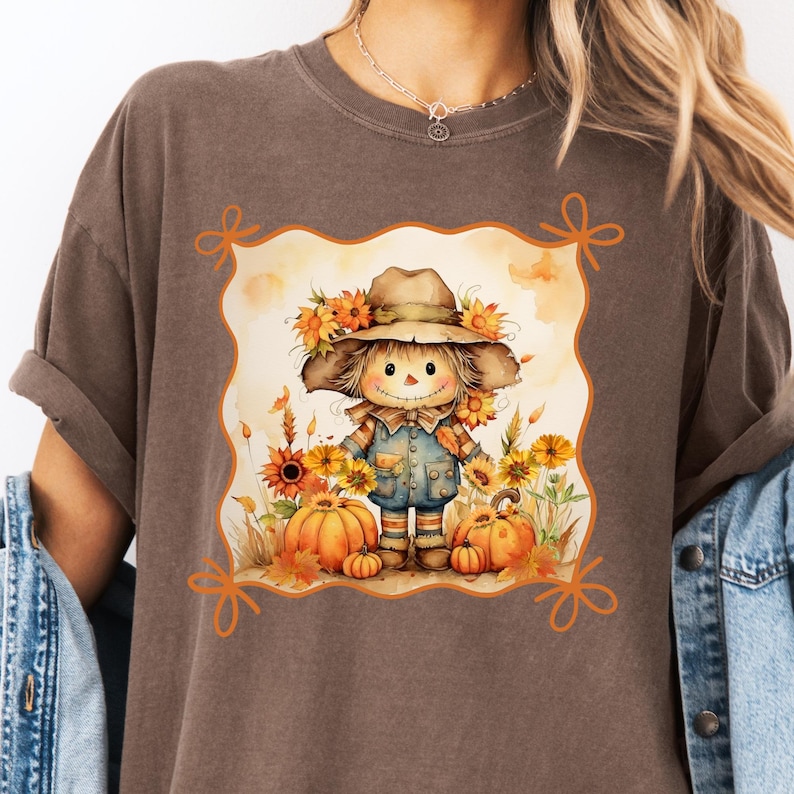 May include: Brown t-shirt featuring a fall-themed graphic. The graphic depicts a smiling scarecrow wearing a hat and overalls, surrounded by pumpkins and sunflowers. The design is framed with orange decorative elements.