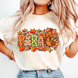 May include: A cream-colored t-shirt featuring a graphic print with the text PARA in large, patterned letters surrounded by autumn leaves, small pumpkins, and a cartoon turkey wearing a plaid bow. The design uses orange, green, and red colors.