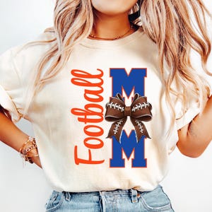 May include: Cream-colored t-shirt with the word "Football" in orange script. The design includes the word "MOM" in blue block letters, with a football-shaped bow in the center. The bow is brown with white laces.