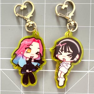 May include: Two anime-style character keychains. One keychain features a character with pink and blue hair, wearing a black outfit. The other keychain has a character with black hair and a white outfit. Both have gold-colored heart-shaped clasps.