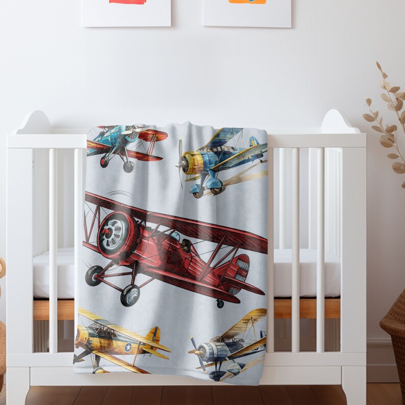 May include: A baby blanket featuring watercolor illustrations of vintage airplanes in shades of red, blue, yellow, and gold. The planes are depicted in flight against a light blue background. The blanket is draped over a white crib.